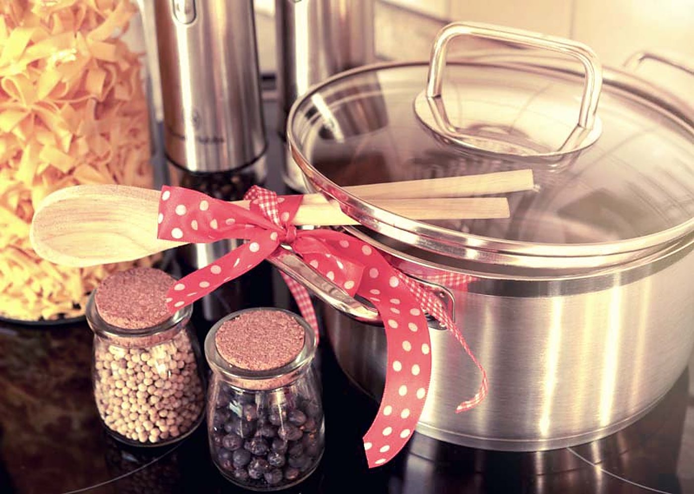 Une casserole en inox à couvercle en verre, une cuillère en bois nouée d'un ruban rouge à pois, deux petits bocaux d'épices et des pâtes sèches sur une plaque de cuisson.