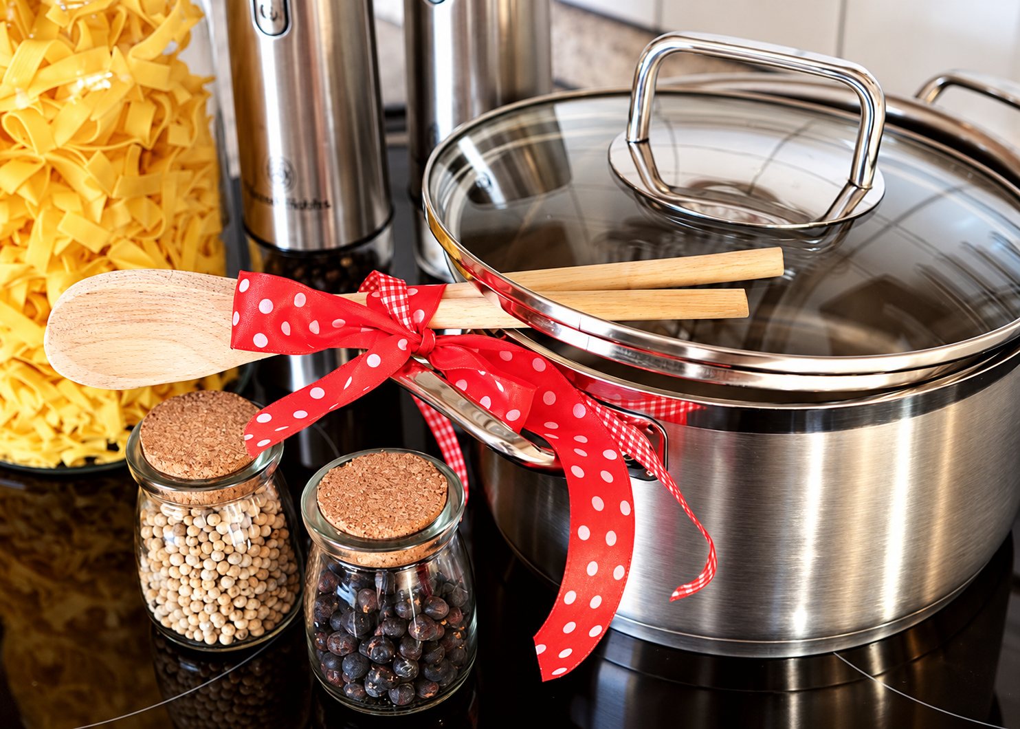 Une casserole en inox à couvercle en verre, nouée d'un ruban rouge à pois avec des cuillères en bois, posée près de deux pots d'épices et de pâtes.