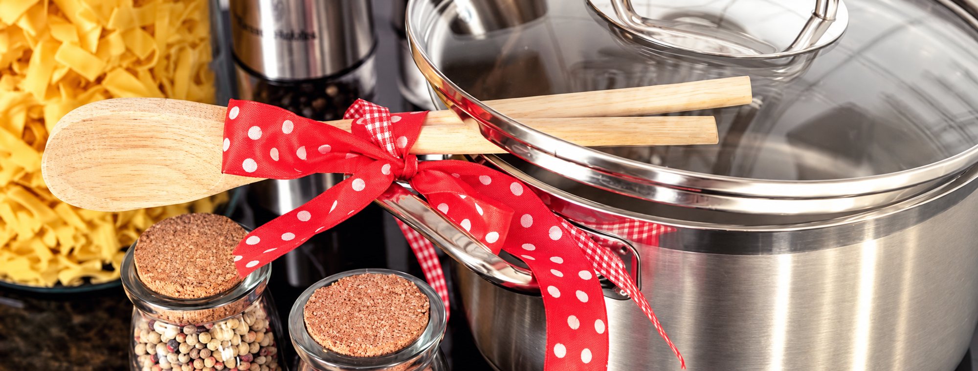 Une casserole en inox à couvercle en verre, ornée d'un ruban rouge à pois blancs qui retient des cuillères en bois, à côté de bocaux d'épices et de pâtes.