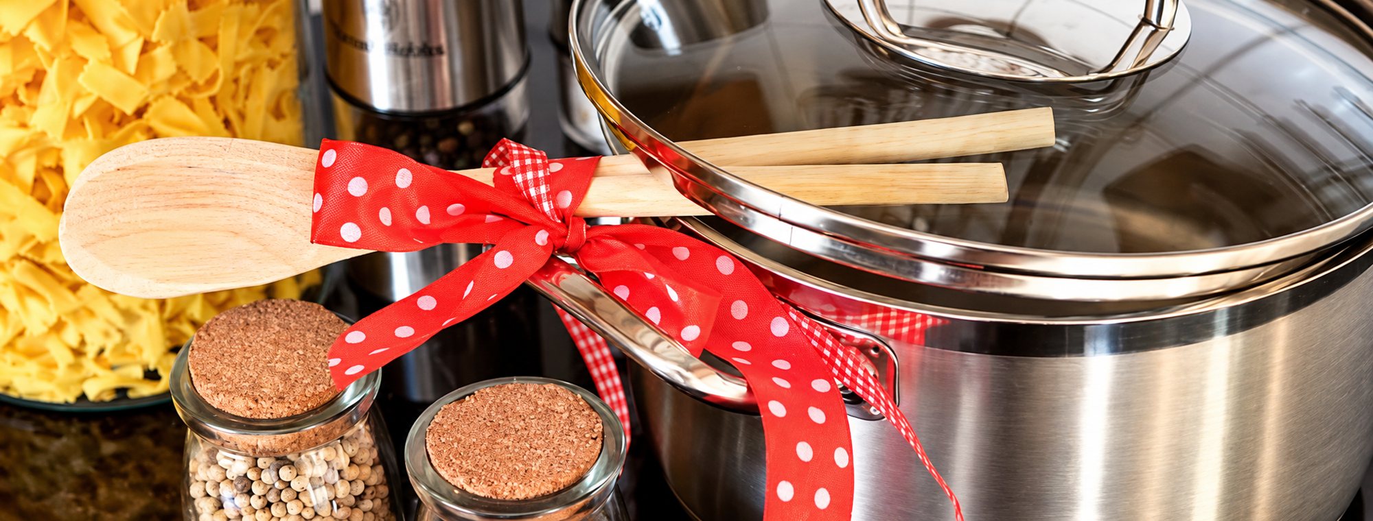 Une casserole en inox avec son couvercle en verre, décorée d'un ruban rouge à pois blancs qui retient des cuillères en bois, à côté de bocaux d'épices et de pâtes.
