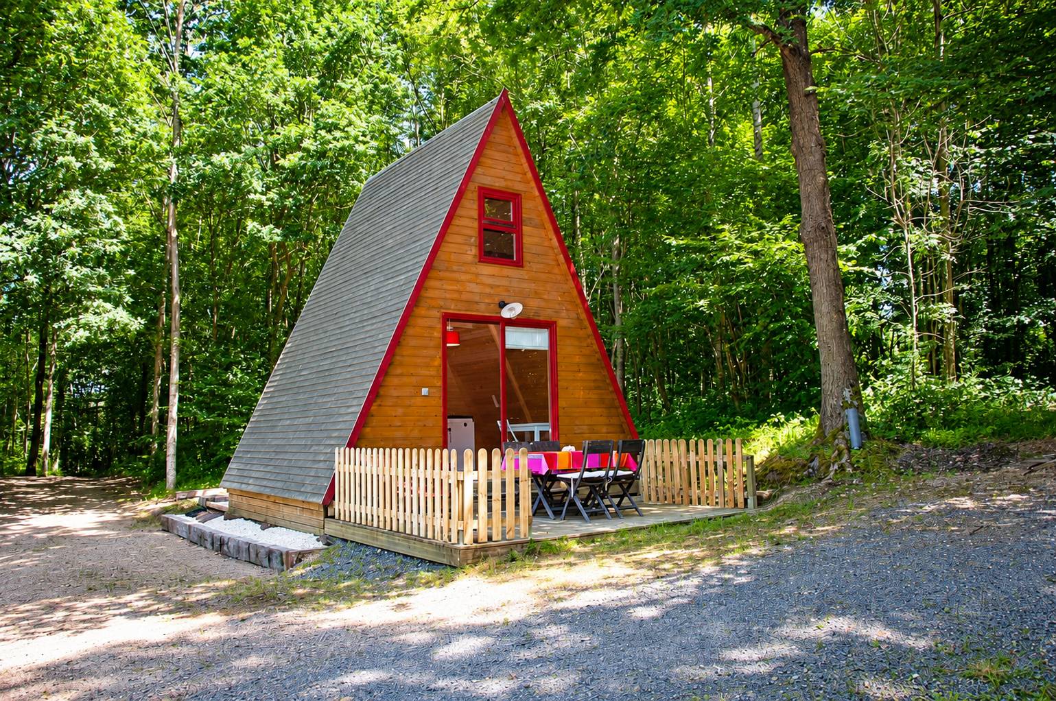 Un petit chalet en bois de forme triangulaire aux bordures rouges, avec une terrasse clôturée et une table dressée, au milieu d'une forêt.