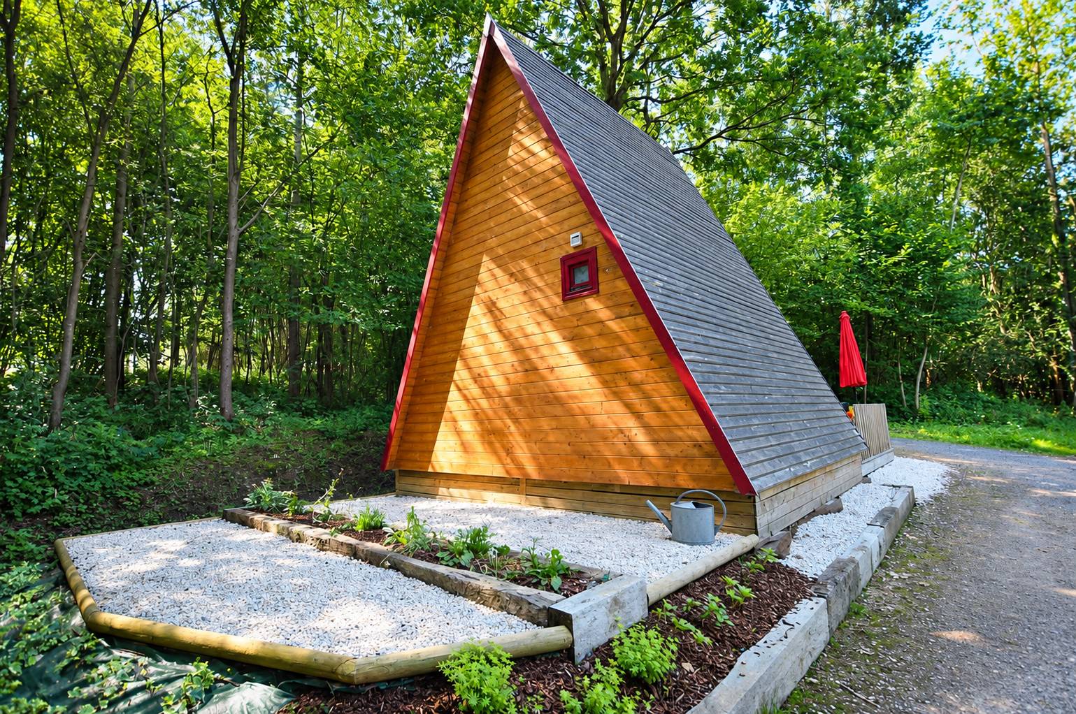 Un chalet en bois en forme de A avec un toit gris et une petite fenêtre rouge, entouré d'arbres, de gravier blanc et de jeunes plantes, avec un arrosoir posé devant.