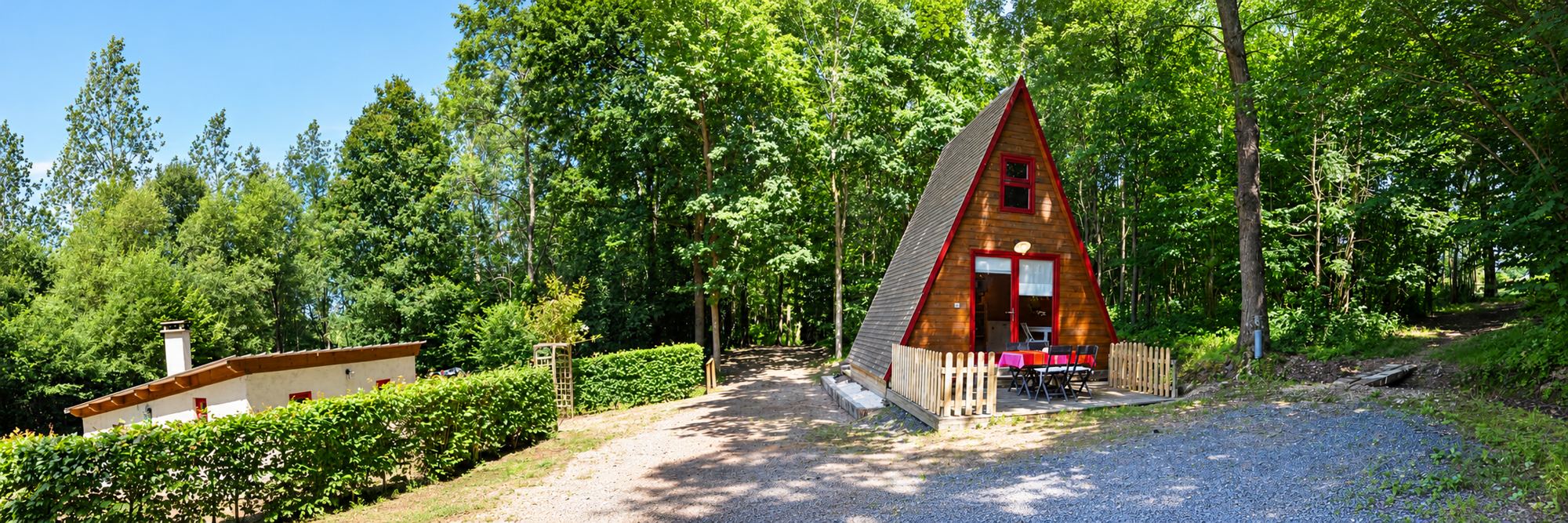 Un chalet en bois en forme de A avec une terrasse et une table rouge, entouré d'arbres et d'une haie, à côté d'un petit bâtiment blanc.