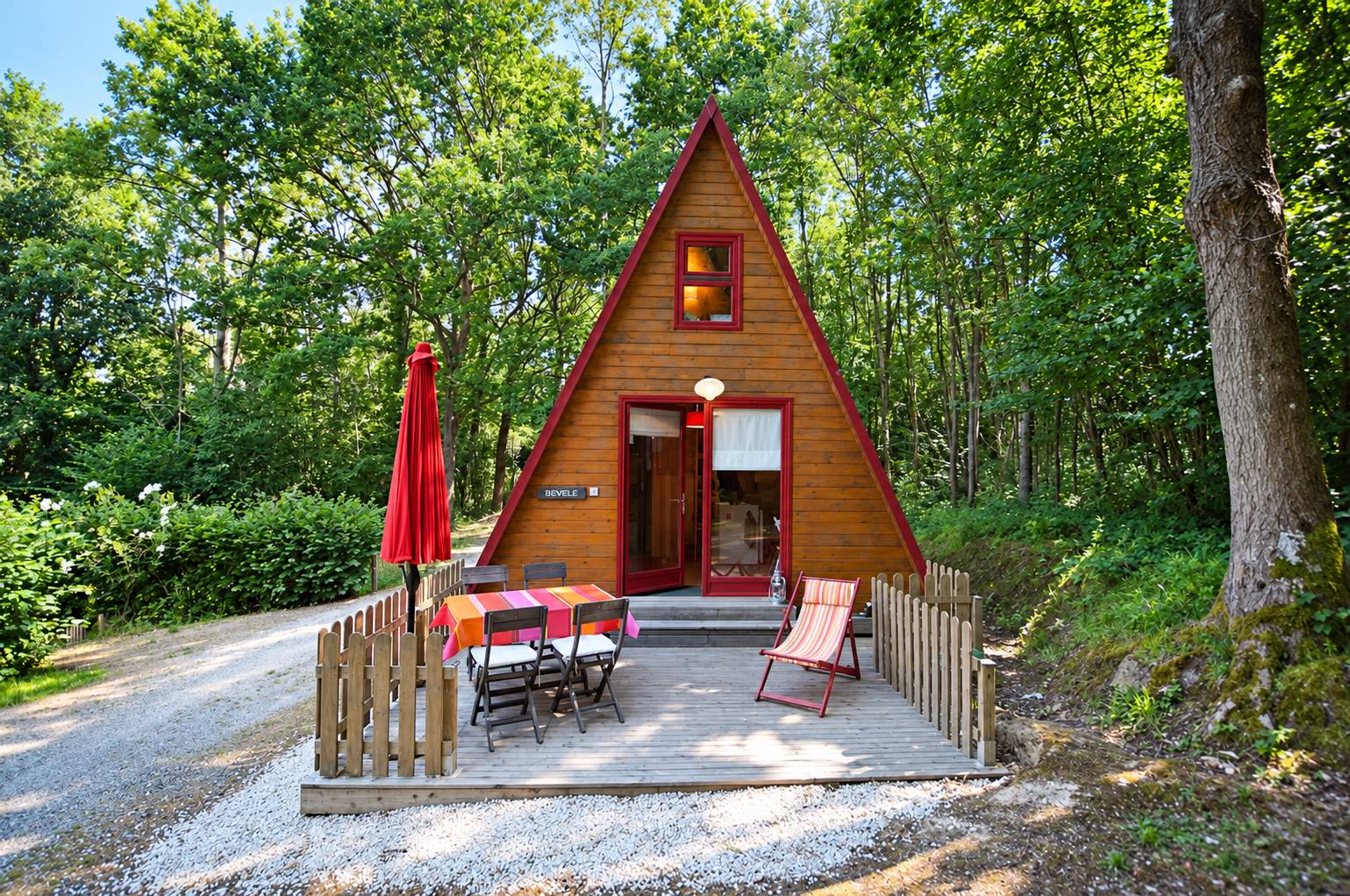 Un petit chalet en bois en forme de A aux bordures rouges, avec une terrasse équipée d'une table, de chaises et d'un parasol rouge, entouré d'arbres.