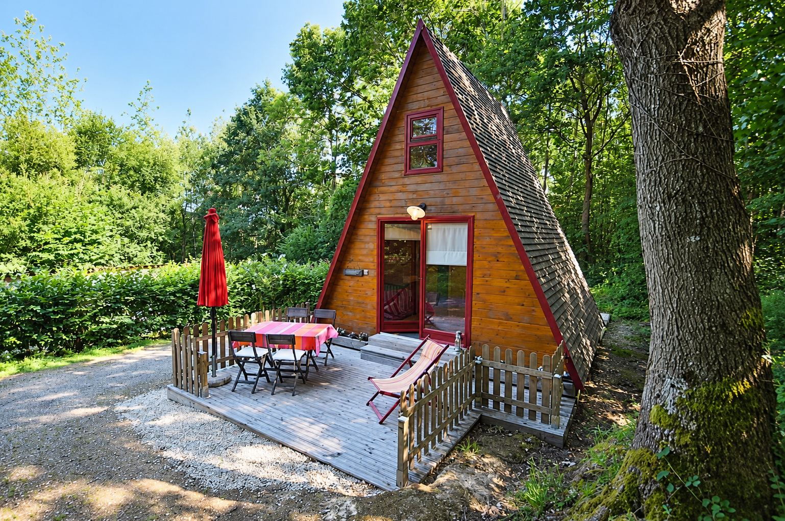 Un chalet en bois en forme de A avec une terrasse équipée d'une table, de chaises et d'un parasol rouge fermé, entouré d'arbres et de verdure.