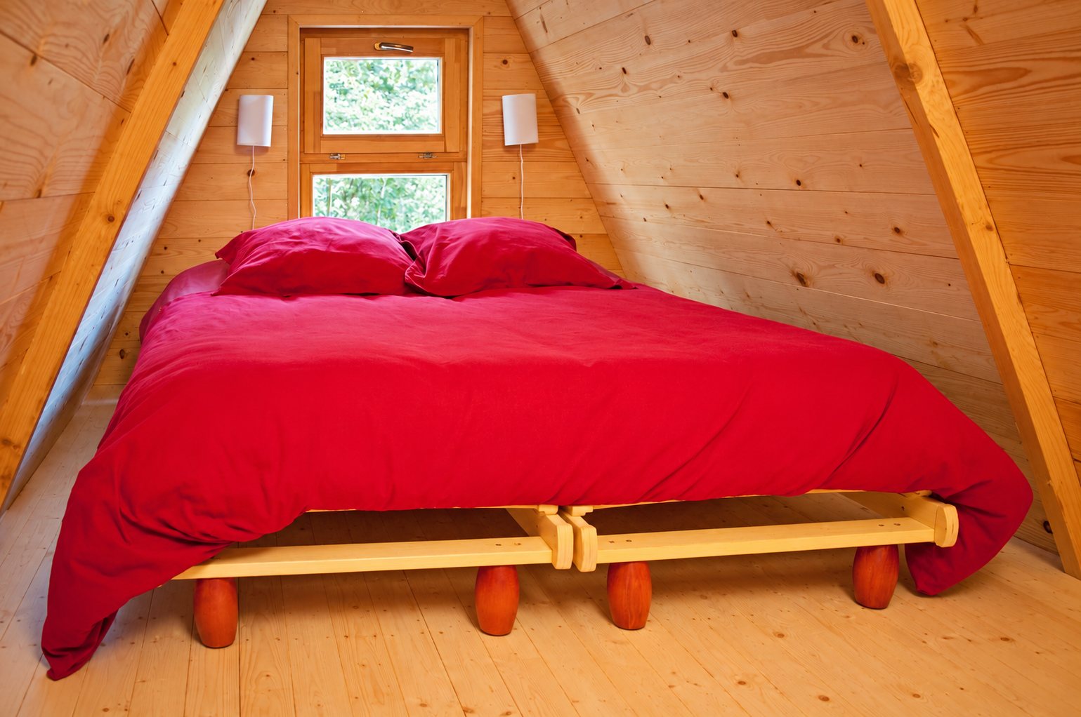 Sous les combles d'un chalet en bois, un grand lit recouvert d'une couette rouge est placé devant une petite fenêtre donnant sur la verdure.