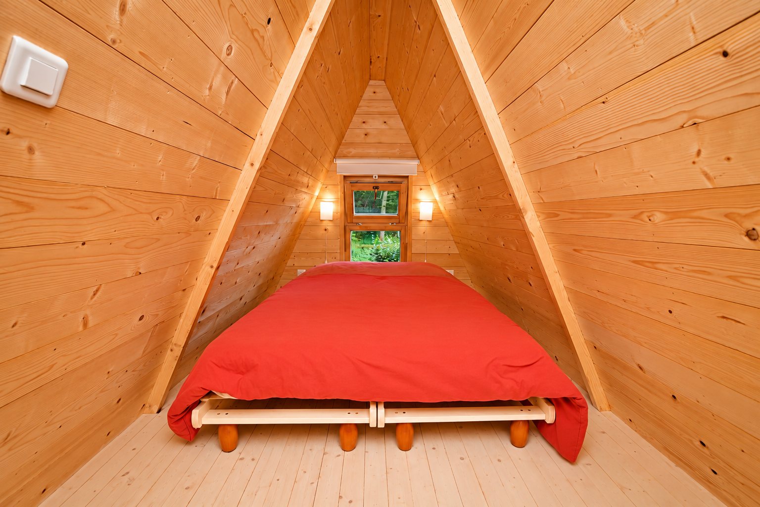 Intérieur d'une petite chambre en bois à toit en pente, avec un lit recouvert d'une couette rouge sous une fenêtre donnant sur la verdure.