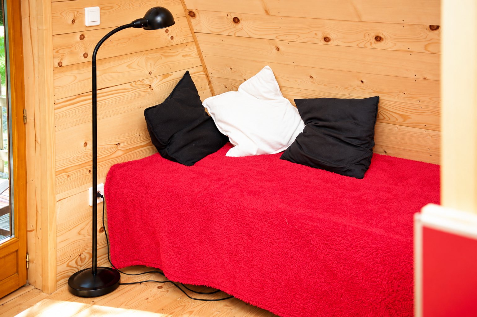 Intérieur d'un hébergement en bois clair avec un lit recouvert d'une couverture rouge, trois coussins noirs et blancs et un lampadaire noir.