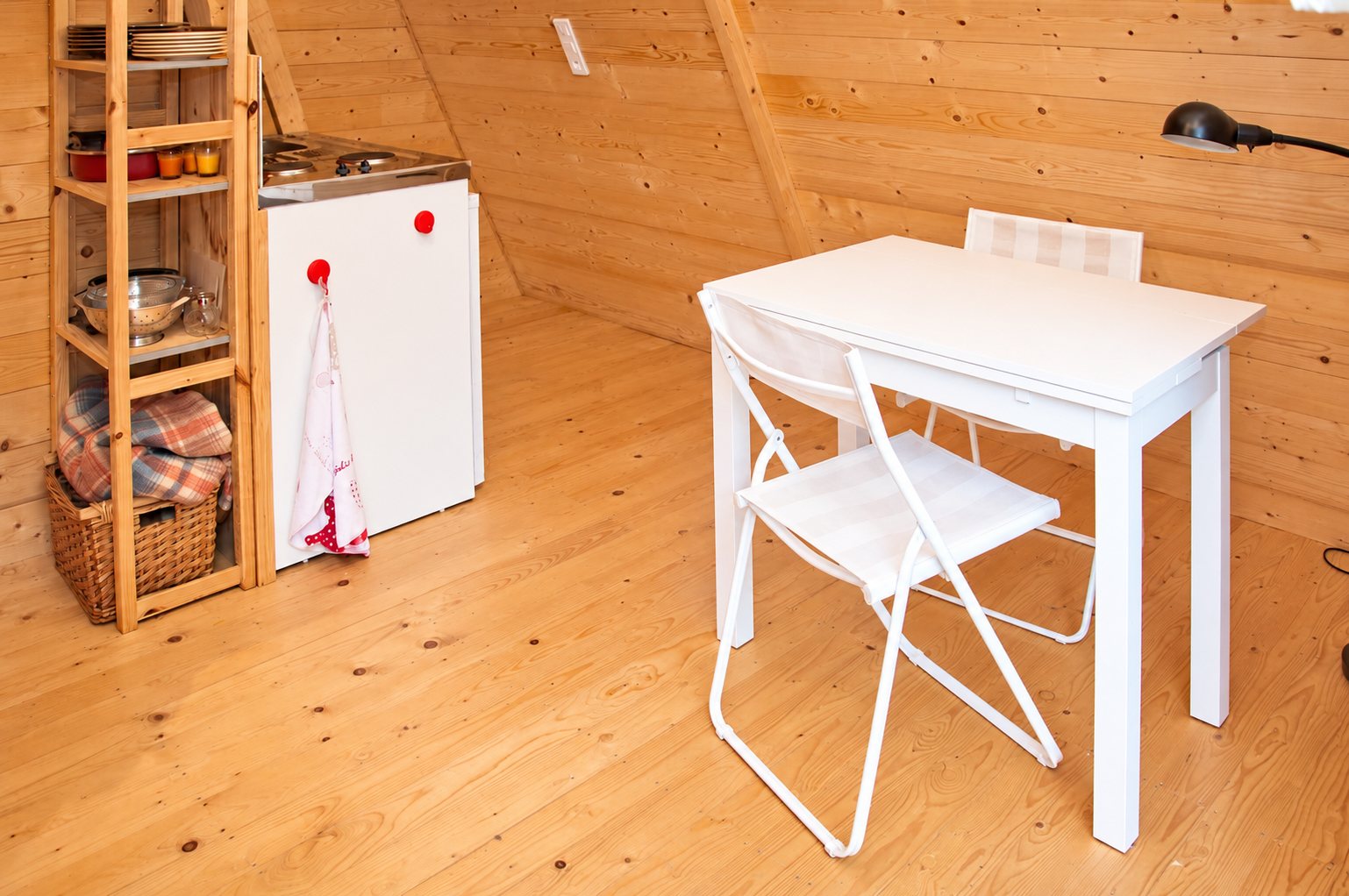 Intérieur d'un petit hébergement en bois avec une table blanche, deux chaises pliantes, une kitchenette et une étagère en bois.