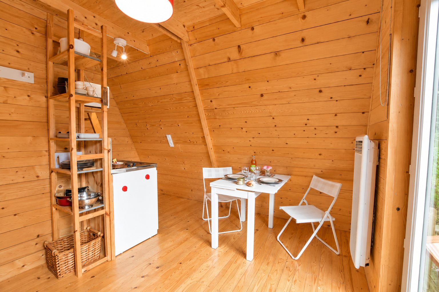 Intérieur lumineux d'un petit chalet en bois avec une table blanche dressée pour deux, deux chaises pliantes, une kitchenette et une étagère en bois.