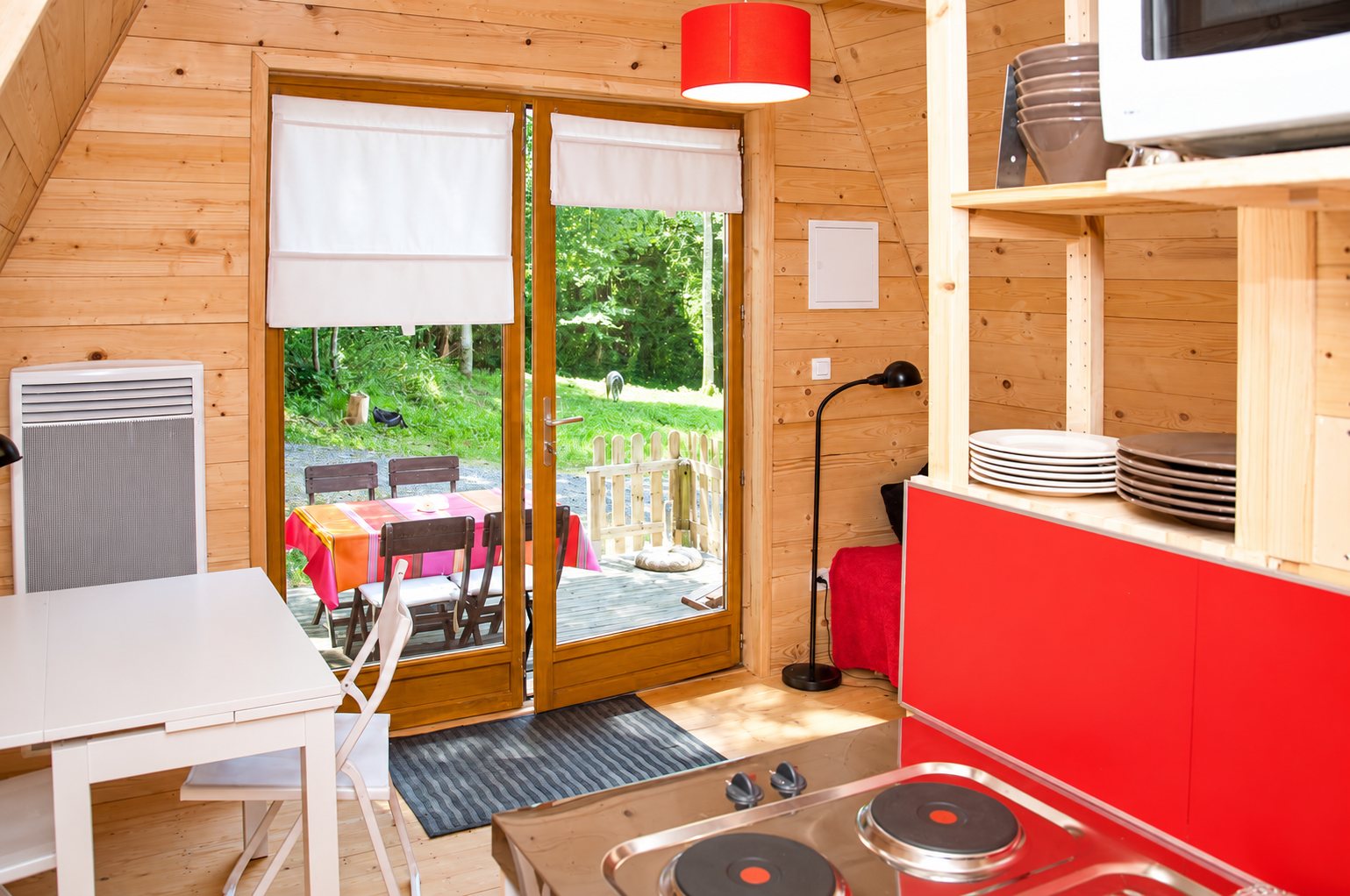 Intérieur lumineux d'un petit chalet en bois avec kitchenette rouge, table blanche et porte-fenêtre donnant sur une terrasse et la forêt.
