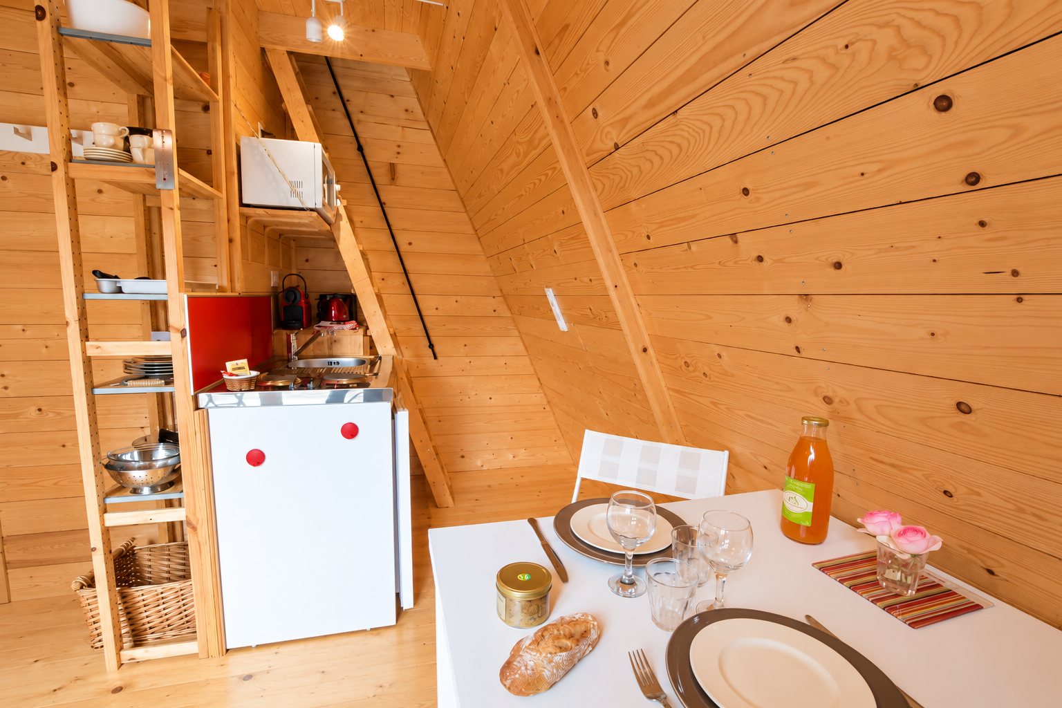 Intérieur d'un petit chalet en bois clair avec une kitchenette, des étagères et une table dressée avec pain, verres et jus d'orange.