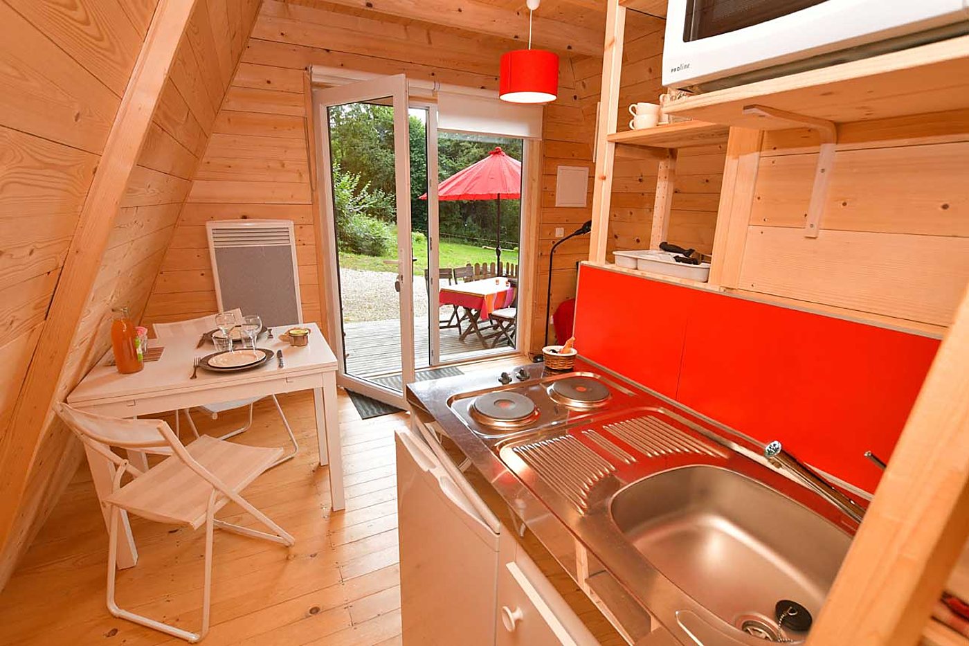 Intérieur d'un petit chalet en bois de forme triangulaire avec une kitchenette rouge, une table dressée pour deux et une porte-fenêtre ouverte sur une terrasse avec parasol rouge.