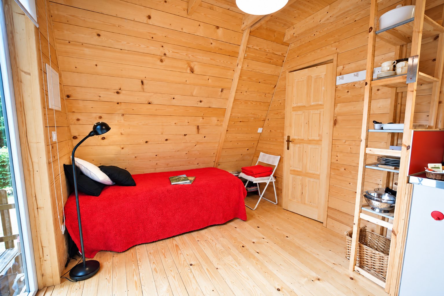 Petite chambre d'un chalet en bois clair avec un lit couvert d'un plaid rouge, une lampe noire, une chaise blanche pliante et des étagères de cuisine.