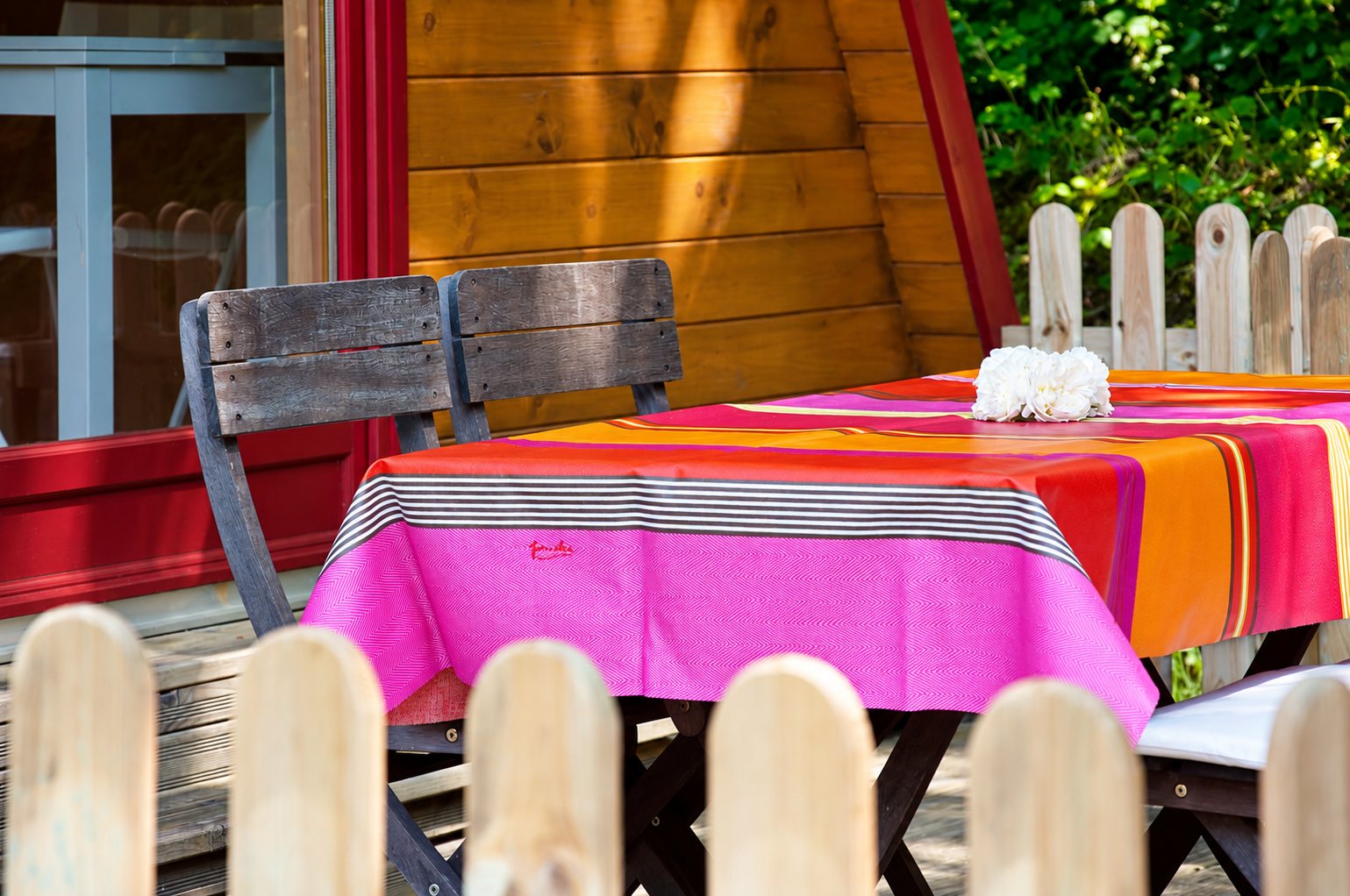 Une nappe rayée rose, orange et rouge recouvre une table de terrasse avec des chaises en bois devant un chalet et une barrière en bois clair.